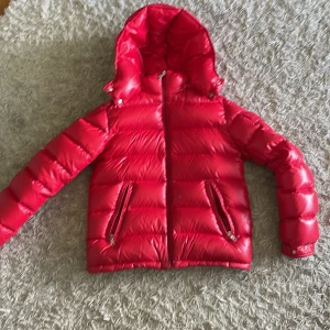 Röd dunjacka från Moncler med huva - Säljer en riktigt snygg röd dunjacka från Moncler med glansig finish och huva. Jackan har dragkedja framtill, två fickor med dragkedja och klassisk Moncler-logga på insidan. Perfekt för kalla dagar när du vill sticka ut.