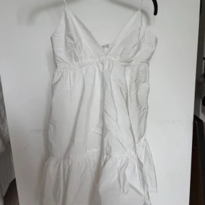 Vit klänning urringad  - Superfin vit kortklänning med smala axelband och v-ringning. Klänningen har en lätt volang nertill och knytning i ryggen. Perfekt för varma dagar och har en luftig passform. Materialet känns som bomull och ger en fräsch vibe.