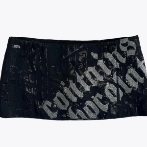 Svart kortkjol från Miss Sixty - Cool svart kortkjol från Miss Sixty med glittrig silvertext i stor gotisk stil över hela framsidan. Kjolen har en rak passform och är tillverkad i ett strukturerat tyg med diskret mönster. Perfekt statement-plagg för dig som vill sticka ut. Kjolen är av omlotttyp i fram, med inbyggd trosa. 25 cm lång.