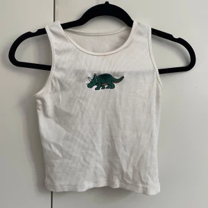 Vit ribbad linnetopp med dinosaurie från Shein - Vit ribbad linnetopp med broderad grön dinosaurie framtill från Shein. Toppen är ärmlös och har en klassisk rund halsringning. Perfekt för dig som gillar coola detaljer och vill sticka ut lite extra.