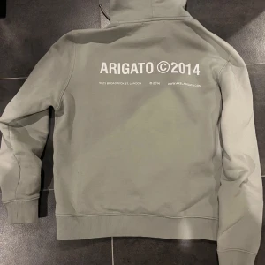 Grå hoodie från Axel Arigato - Säljer en stilren grå hoodie från Axel Arigato med tryck 'ARIGATO ©2014' på både bröstet och ryggen. Tröjan har huva med dragsko och en stor magficka. Perfekt för en avslappnad look. Hoodien är i nyskick och produceras inte längre. 
