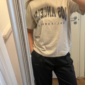 Beige Los Angeles t-shirt - Beige t-shirt med svart tryck där det står 'Los Angeles California' i college-stil. T-shirten har klassisk passform, rund hals och korta ärmar. Perfekt basic med coolt print som funkar till allt.