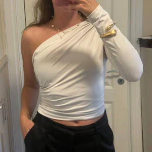 Ljusbeige one shoulder topp i stretch - Säljer en ljusbeige, figurnära one shoulder topp med lång ärm på ena sidan. Toppens material är stretchigt och slätt, vilket ger en snygg draperad effekt över kroppen. Perfekt för dig som gillar en stilren och trendig look.