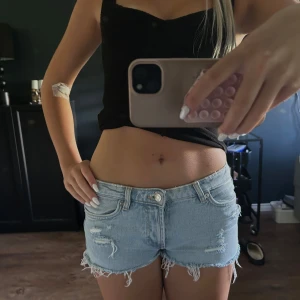Ljusblå jeansshorts med slitningar - Säljer ett par ljusblå jeansshorts med råa kanter och slitna detaljer framtill. Shortsen har låg midja, klassiska fickor och bälteshällor. Perfekta för varma dagar när du vill ha en chill och avslappnad look.