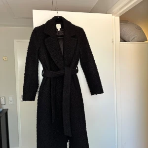 Svart teddyrock från H&M med bälte - Säljer en svart lång teddyrock från H&M i storlek 36. Jackan har en mysig, fluffig struktur, klassisk krage och knyts med bälte i midjan. Perfekt för kalla dagar när du vill vara både varm och snygg. Långärmad och stilren design.