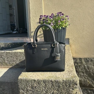 Svart handväska från Don Donna - Stilren svart handväska från Don Donna med guldfärgade detaljer och två rejäla handtag. Väskan har en klassisk, boxig form och är tillverkad i ett strukturerat syntetmaterial. Invändigt finns flera fack och dragkedja för smidig organisering. Perfekt för dig som vill ha en snygg och praktisk väska. Orginalpris 499kr! Är använd i 1 vecka.