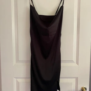 Svart satinklänning med slits XS - Svart satinklänning i storlek XS med tunna axelband och snygg draperad urringning. Klänningen har en liten slits på ena sidan