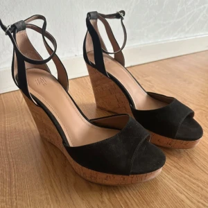 Svarta kilklackade sandalery H&M - Snygga svarta sandaler från H&M med hög kilklack i kork och öppen tå. Ovandelen är i mocka-look och de har smala remmar med spänne runt ankeln. Perfekta för sommarens alla outfits och ger en cool och trendig vibe