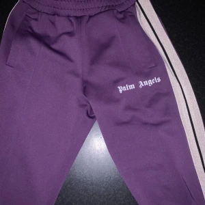 Palm Angels byxor  - Snygga lila mjukisbyxor från Palm Angels med vita och svarta revärer längs sidorna. Byxorna har elastisk midja, raka ben och logga tryckt på ena låret. Tillverkade i 100% polyester för en sportig och bekväm känsla.