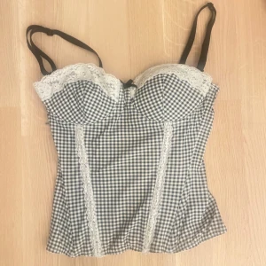 Rutig korsett med spets - Supersöt korsett med svartvitt rutigt mönster med vita spetsdetaljer längs kuporna. Från H&M som ej säljs längre i storlek s/36.Justerbara svarta axelband och hakknäppning bak. Super gullig men passar tyvärr inte mig längre:/ Inte använt så många gånger och har inga synliga defekter.