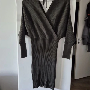 Mörkgrå ribbad omlottklänning - Snygg mörkgrå ribbad klänning med omlott överdel och v-ringning. Klänningen har långa ärmar och knytband i nacken. Den är figurnära nertill och har en cool struktur med bredare ribbor upptill och smalare nedtill. Perfekt för dig som gillar stilrena och trendiga plagg.