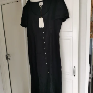 Svart långklänning i linne från KL o co  - Stilren svart långklänning från KL o co i svalt linne. Klänningen har korta ärmar, rundad urringning och ljisa knappar längs hela framsidan. Perfekt för dig som gillar minimalistisk och tidlös design. Aldrig använd helt ny med prislappen kvar. Fick inte byta den för det gått för lång tid. Inköpt i butiken i K Lagerqvist i Varberg. Superfin o skön men på mig är den för tajt över bysten. Så därför säljer jag den vidare. Nypris 1900kr 