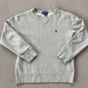 Grå sweatshirt från Polo Ralph Lauren - Klassisk grå sweatshirt från Polo Ralph Lauren med rund halsringning och broderad marinblå logga på bröstet. Tröjan har långa ärmar, ribbade muddar och är tillverkad i mjuk bomullsmix. Perfekt för en chill och stilren look.