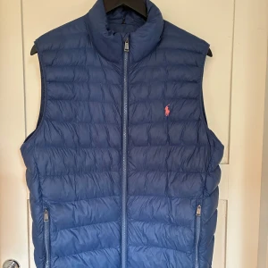 Blå dunväst från Ralph Lauren - Snygg blå dunväst från Ralph Lauren med quiltad design och hög krage. Västen har dragkedja framtill, två fickor med dragkedja och den klassiska rosa Polo-loggan broderad på bröstet. Perfekt att slänga över en tröja eller skjorta nu i sommar för extra värme. Västen går lätt att packa ned i innerfickan för att ha med den på semestern eller väskan någonstans. Nypris 2590kr, mitt pris 1000kr.