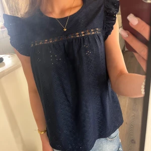 Mörkblå blus med volang från SHEIN - Supersöt mörkblå blus från SHEIN i storlek XS. Blusen har broderade detaljer, volangärmar och ett fint hålmönster över hela tyget. Ovanför bysten finns en dekorativ spetskant med små blommor. Perfekt till jeans eller kjol för en söt look.