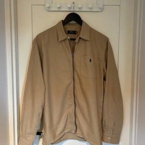 Beige overshirt från Polo Ralph Lauren - Snygg beige overshirt från Polo Ralph Lauren med dragkedja framtill och klassisk krage. Bröstficka med broderad logga i mörkblått. Långärmad modell i bomull, perfekt att slänga över en t-shirt eller skjorta nu i sommar. Nypris 1900, mitt pris 850