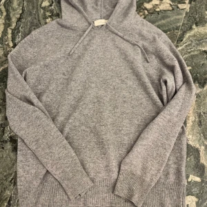 Grå kashmir hoodie - Mysig grå hoodie från Soft Goat i stickad kashmir. Tröjan har huva med snörning, långa ärmar och breda ribbade muddar vid ärmslut och nederkant. Perfekt för dig som gillar en clean och chill stil.
