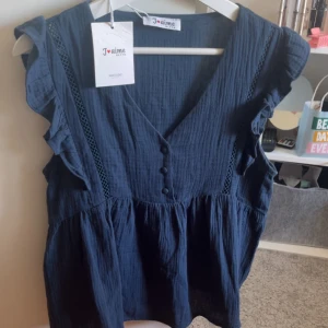 Mörkblå blus med volang från missäy  - Somrig mörkblå blus från missäy med volangdetaljer på axlarna, v-ringning och dekorativa knappar framtill. Blusen har ärmlös design och är gjord i mjuk bomull med fina spetsinslag längs axlarna. Perfekt för dig som gillar en romantisk och luftig stil. Aldrig använt och ny med prislapp på.