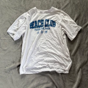 Vit BEACH CLUB t-shirt från Lager 157 - Vit t-shirt från Lager 157 med blå BEACH CLUB-tryck och hibiskusblommor framtill. Klassisk rund halsringning och korta ärmar. Tillverkad i 100% bomull, perfekt för chill dagar och sommarvibbar.