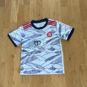 FC Bayern München Adidas fotbollströja - Snygg FC Bayern München fotbollströja från Adidas med vit bas och mörkblå grafiskt mönster. Röd klubbmärke på bröstet och röda Adidas-ränder på axlarna. Svart ribbad krage och ärmslut. Tillverkad i lätt och ventilerande material, perfekt för match eller träning.
