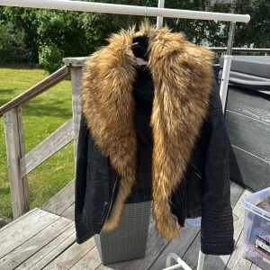 Svart skinnjacka med fuskpäls från Chica London - Cool svart skinnjacka från Chica London i storlek S med stor, fluffig fuskpäls i bruna toner runt kragen. Jackan har snygga detaljer med dragkedjor och quiltade partier på ärmarna. Perfekt statement-plagg för dig som vill sticka ut.
