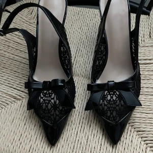 Svarta spetsiga pumps med rosett - Snygga svarta pumps med spetsig tå och dekorativ rosett framtill. Skorna har detaljer i blankt material och sidor i spets, vilket ger en elegant look. Perfekta för dig som gillar stilrena och trendiga skor med det lilla extra.