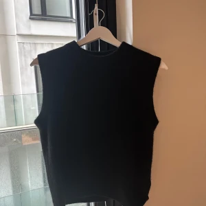 Svart stickad väst i XS från ARKET - En svart, ärmlös stickad väst med rund halsringning. Västen har en enkel och stilren design utan synliga detaljer eller mönster, vilket gör den lätt att matcha med olika outfits. Perfekt för lager-på-lager och passar dig som gillar minimalistisk stil.