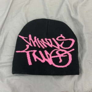 Svart mössa med rosa graffiti-tryck - Cool svart mössa med ett stort rosa graffiti-inspirerat tryck framtill. Mössan är stickad och har en klassisk form som sitter snyggt på huvudet. Perfekt accessoar för att sticka ut i mängden och ge din outfit en streetstyle vibe.