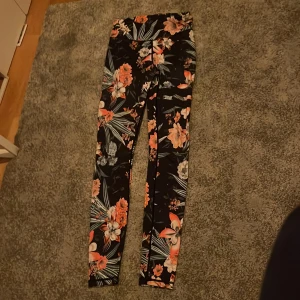Blommiga svarta leggings - Svarta leggings från Stronger med blommiga detaljer, passar perfekt för en springtur, jag är osäker på storlek men tror det är S eller M . Passar mig relativt bra i längden och jag är 161cm. Skulle inte säga att de är slitna förutom att stronger namnet har slitits bort för mesta del, annars skulle jag bara säga att de är använda. Ge ett pris!!