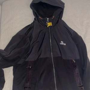 Svart vindjacka från Parajumpers - Snygg svart vindjacka från Parajumpers med huva och hel dragkedja framtill. Jackan har logga på bröstet, praktiska fickor och en gul detalj vid halsen. Perfekt för dig som vill ha en stilren och funktionell jacka till vardags. Den är äkta lite solbränd