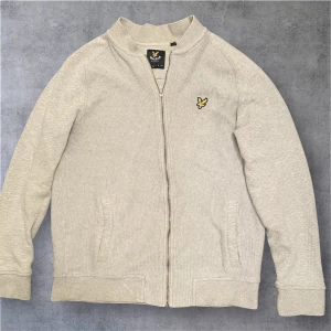 Beige cardigan jacka/tröja från Lyle & Scott - Snygg beige cardigan jacka från Lyle & Scott med dragkedja framtill och broderad gul logga på bröstet. Jackan är stickad i bomull och har ribbade muddar samt två fickor. Perfekt för dig som gillar stilren och enkel design.
