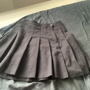 Svart kjol  - Svart kort plisserad kjol från Divided i storlek 36. Kjolen har bred linning med knapp och klassiska djupa veck som ger en snygg volym. Perfekt för dig som gillar en stilren och edgy look.