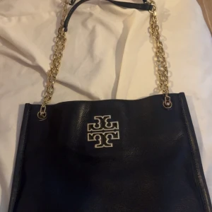 Svart axelväska från Tory Burch - Snygg svart axelväska från Tory Burch i äkta skinn med guldfärgade kedjor som axelrem. Väskan har en stilren design med det klassiska Tory Burch-emblemet i silver på framsidan. Perfekt storlek för det viktigaste och ger en lyxig känsla till din outfit.