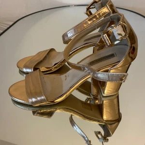 Guldiga sandaler med klack - Snygga sandaler i metallic guld från Bianco med bred rem över tårna och justerbar rem runt vristen. Skorna har öppen tå och blockklack som ger stabilitet. Perfekta för att lyfta din outfit med en glansig touch.