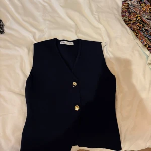 Mörkblå stickad väst från Zara - Snygg mörkblå stickad väst från Zara med v-ringning och två guldiga knappar framtill. Västen har en klassisk, rak passform och är perfekt att styla över en skjorta eller t-shirt för en clean look.