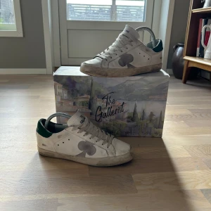 Vita sneakers med grön häl, the Gallerist - Säljer ett par vita sneakers från The Gallerist med gröna detaljer på hälen och en stor grå klöver på sidan. Skorna har snörning, rund tå och platt sula. Tillverkade i skinn och passar perfekt för en avslappnad stil.
