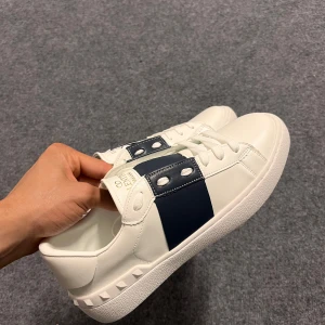 Valentino Garavani vita sneakers - Snygga vita sneakers från Valentino Garavani med bred mörkblå rand och coola nitar på sulan. Skorna har snörning och är tillverkade i skinn för en lyxig känsla. Perfekta för dig som vill sticka ut med stilrena detaljer och exklusiv design.