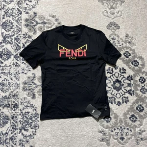 Svart Fendi t-shirt med tryck - Svart t-shirt från Fendi med ikoniskt gult och rött tryck framtill. Klassisk rund halsringning och korta ärmar. Tillverkad i mjuk bomull och har Fendi-tag på sidan. Perfekt för dig som gillar exklusiva streetwear-vibbar.
