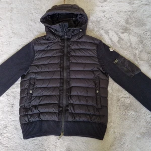 Svart Moncler pufferjacka med huva - Snygg svart pufferjacka från Moncler med stickade ärmar och quiltad framsida. Jackan har huva, dragkedja, ribbade muddar och två fickor med tryckknappar. Perfekt för kyliga dagar och har en clean, sportig vibe. Jackan är som ny, inköpt på NK i januari-25 för 11999 kronor men bara använd 2 ggr. Kvitto samt tags (dock lösa) finns kvar.