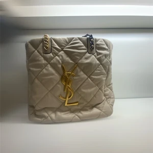 Beige quiltad axelväska  - Säljer en beige quiltad axelväska med stora guldfärgade logodetaljer och kedjeremmar i både guld och svart. Väskan har en lyxig känsla med mjukt skinn och klassiskt rutmönster. Perfekt för dig som vill sticka ut med en ikonisk designerbag.