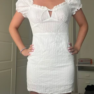 Vit kort klänning med puffärm - Supersöt vit kortklänning med puffärmar och söt knytning framtill. Klänningen har en lätt rynkad struktur och volang vid urringningen.