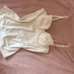 Vit linnetopp med spets från Bon'A Parte - Supersöt vit linnetopp från Bon'A Parte med tunna justerbara axelband och dekorativ spetskant vid kuporna. Toppen har en feminin form med lätt vadderade kupor och små rosetter på banden. Perfekt att styla med jeans eller kjol för en fräsch look.