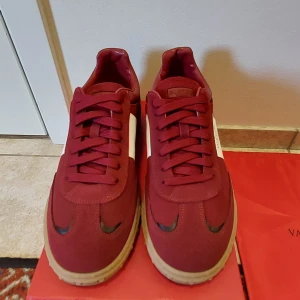 Valentino Garavani röda sneakers - Snygga röda Valentino Garavani sneakers med vita sidopaneler och beige sula. Skorna har klassisk snörning, rund tå och är tillverkade i en mix av skinn och textil. Perfekta för dig som vill sticka ut med stilrena och exklusiva sneakers.