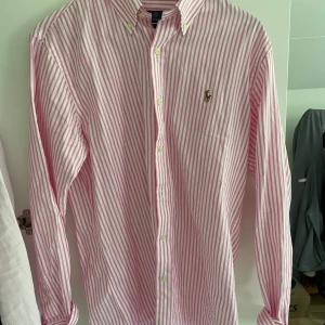 Rosa och vit randig skjorta Polo Ralph Lauren - Snygg rosa och vit randig skjorta från Polo Ralph Lauren i custom fit. Skjortan har klassisk krage, knappar hela vägen och broderad logga på bröstet. Tillverkad i mjuk bomull och har långa ärmar. Perfekt för en clean och fräsch look.