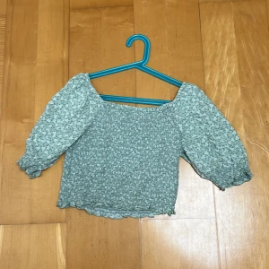 Grön blommig blus med puffärmar - Supersöt ljusgrön blus med småblommigt mönster och puffiga trekvartsärmar. Smockad kropp ger en tight passform och volangkant nertill. Perfekt för dig som gillar romantisk och trendig stil.