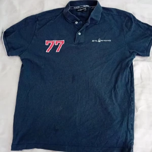 Mörkblå pikétröja Sail Racing - Snygg mörkblå pikétröja från Sail Racing med klassisk krage och två knappar. Framsidan har broderad logga och stort rött 77-tryck. Tröjan är i bomull och har korta ärmar med vita detaljer vid ärmslut. Perfekt för en sportig och avslappnad stil.