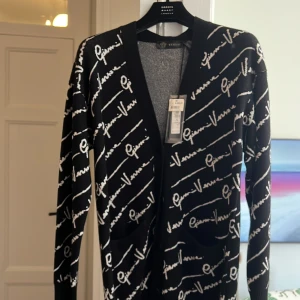 Svart kofta från Versace med logomönster - Svart kofta från Versace med vit logotyptext över hela plagget. Långärmad och öppen framtill med ribbade muddar och två fickor. Snygg och trendig design som sticker ut, perfekt för dig som gillar lyxiga statement-plagg.