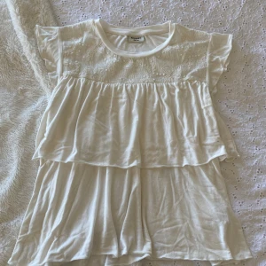 Vit volangtopp från Kappahl - Supersöt vit topp från Kappahl med volanger och broderad överdel. Toppen har korta ärmar och är gjord i mjukt bomullstyg. Perfekt för sommarens varma dagar och har en luftig, flowy passform. Hör av er vid frågor!🥰💞