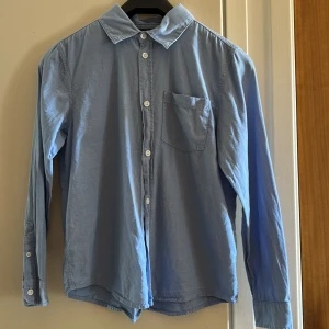 Ljusblå skjorta från Lindex, strl 152 - Klassisk ljusblå skjorta från Lindex i storlek 152. Skjortan har lång ärm, bröstficka och vita knappar framtill samt vid ärmslut. passar perfekt till en avslappnad men ändå stilren look.