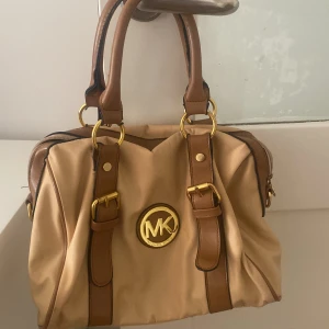 Beige handväska från Michael Kors - Snygg beige handväska från Michael Kors med bruna skinnhandtag och detaljer i guld. Väskan har ett stort MK-emblem framtill och dragkedja upptill. Insidan är fodrad med svart tyg med MK-logga. Perfekt storlek för allt du behöver ha med dig.
